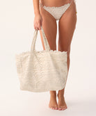 A woman holding a tan lace tote with scalloped trim against a white wall.
