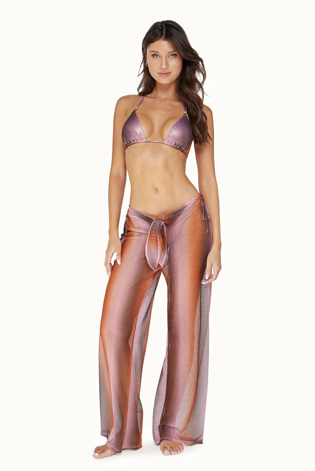 A brunette woman wearing a purple bikini top and long purple pants standing against a white wall.