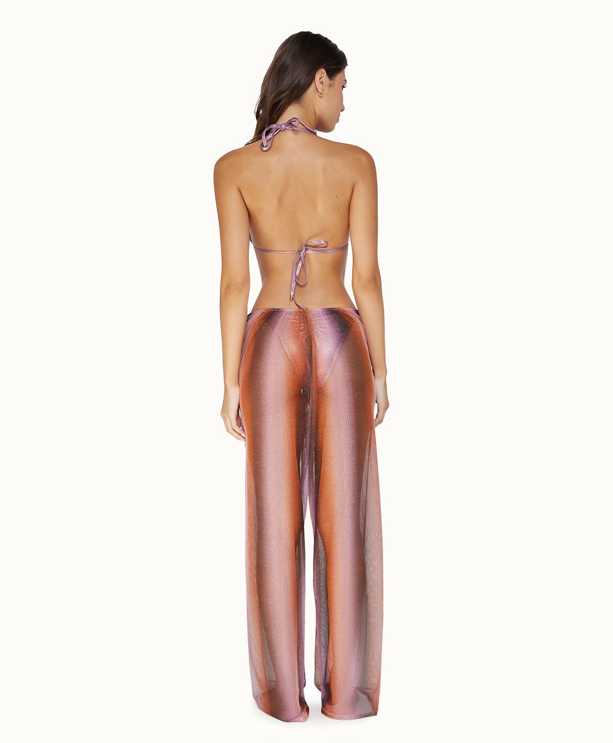 A brunette woman wearing purple pants facing towards a white wall.