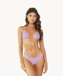 A brunette woman wearing a violet bikini standing in front of a white wall.