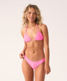 A woman wearing a pink triangle top and bikini bottom standing against a white wall.