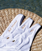 Three white bikini bottoms with gold embroidering laying flat on a wicker basket near the water.
