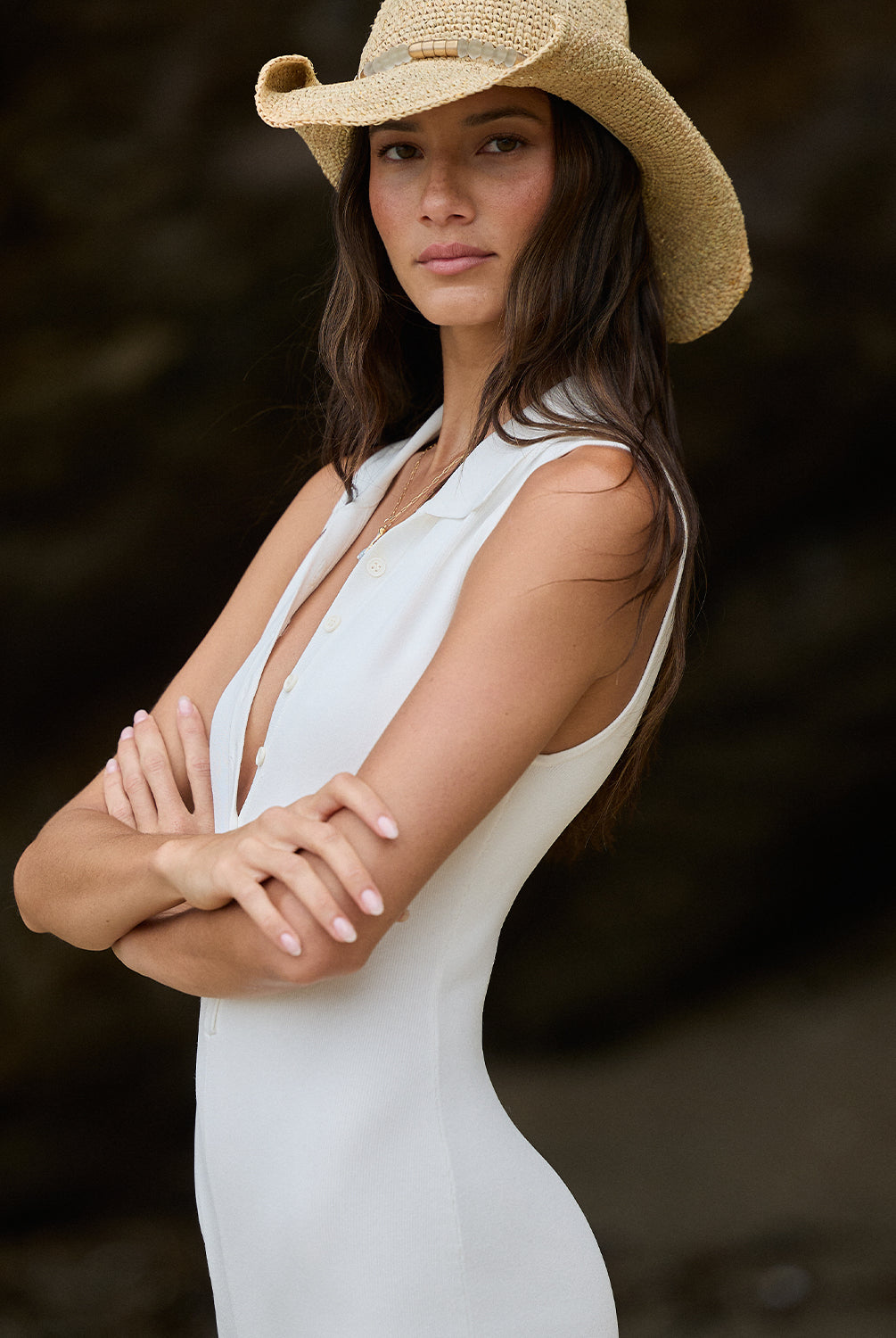 A woman wearing a white romper and cowboy hat standing outside near a cave.