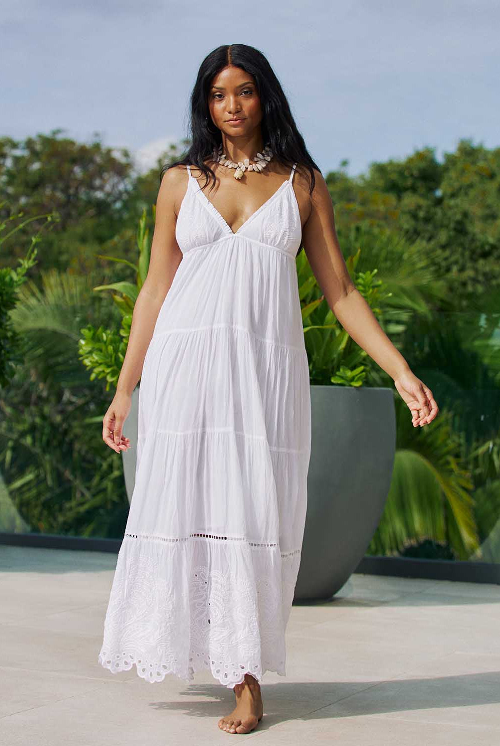 A woman wearing a long white dress withe embroidery details standing outside near plants.