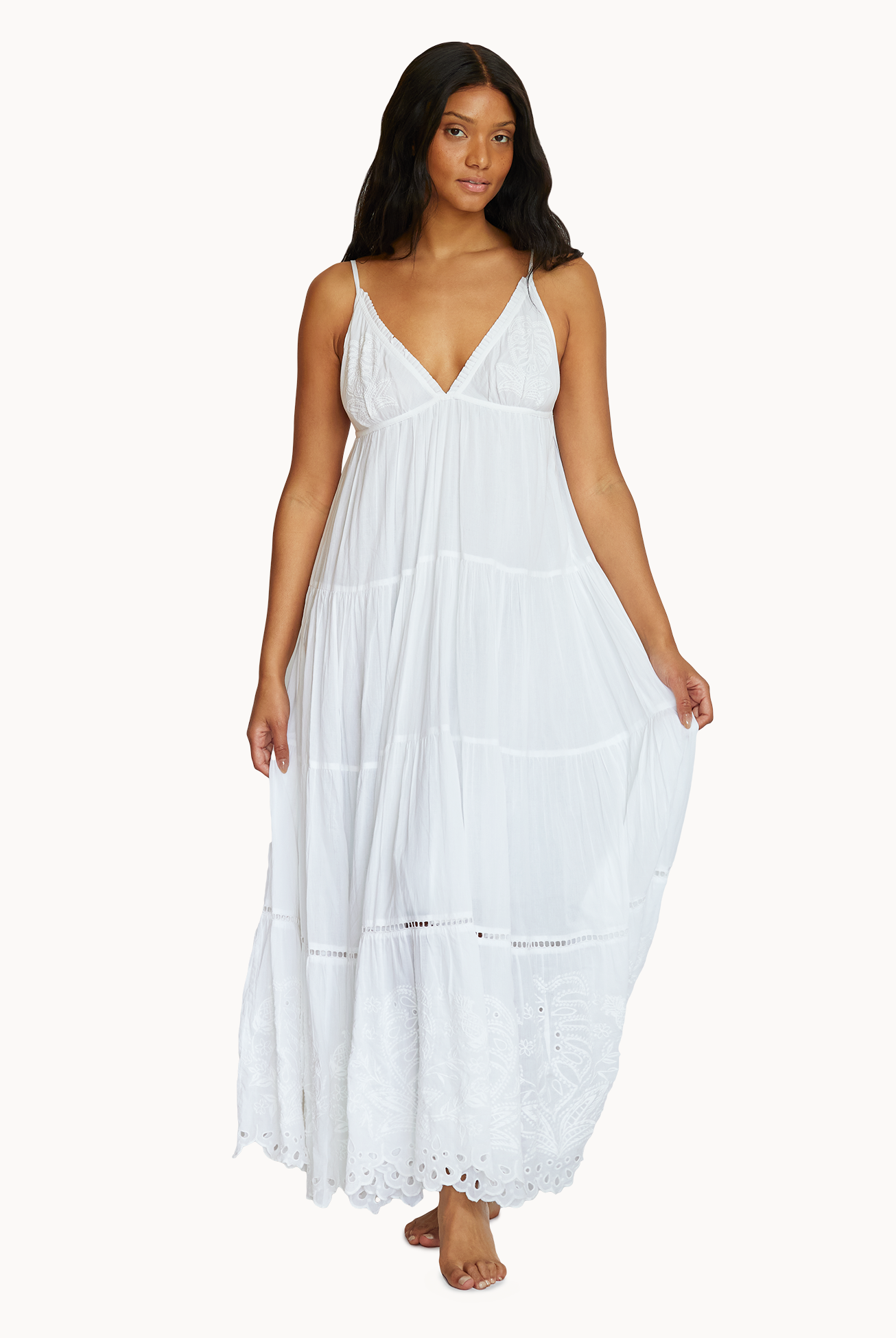 A woman wearing a long white dress with detailed embroidery standing against a white wall.