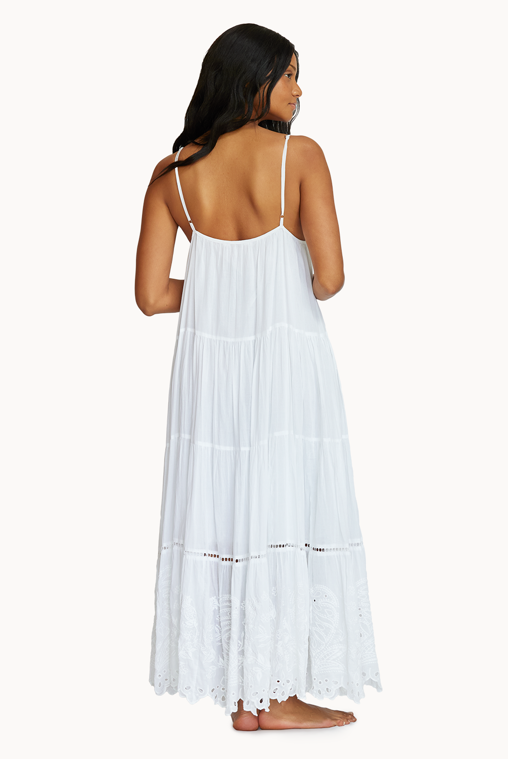 A woman wearing a long white dress with detailed embroidery facing towards a white wall.