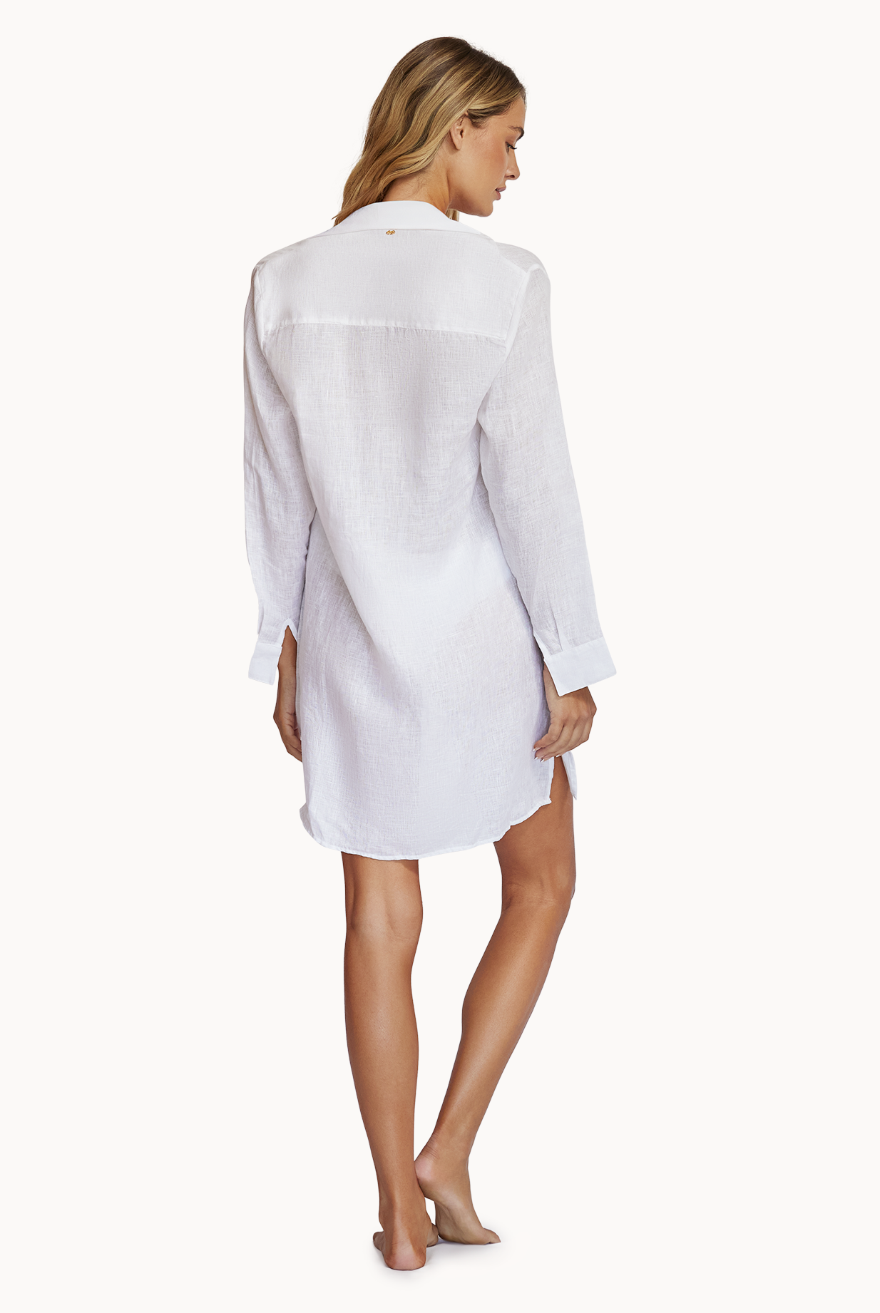 A woman wearing a white long sleeve cover up facing towards a white wall.