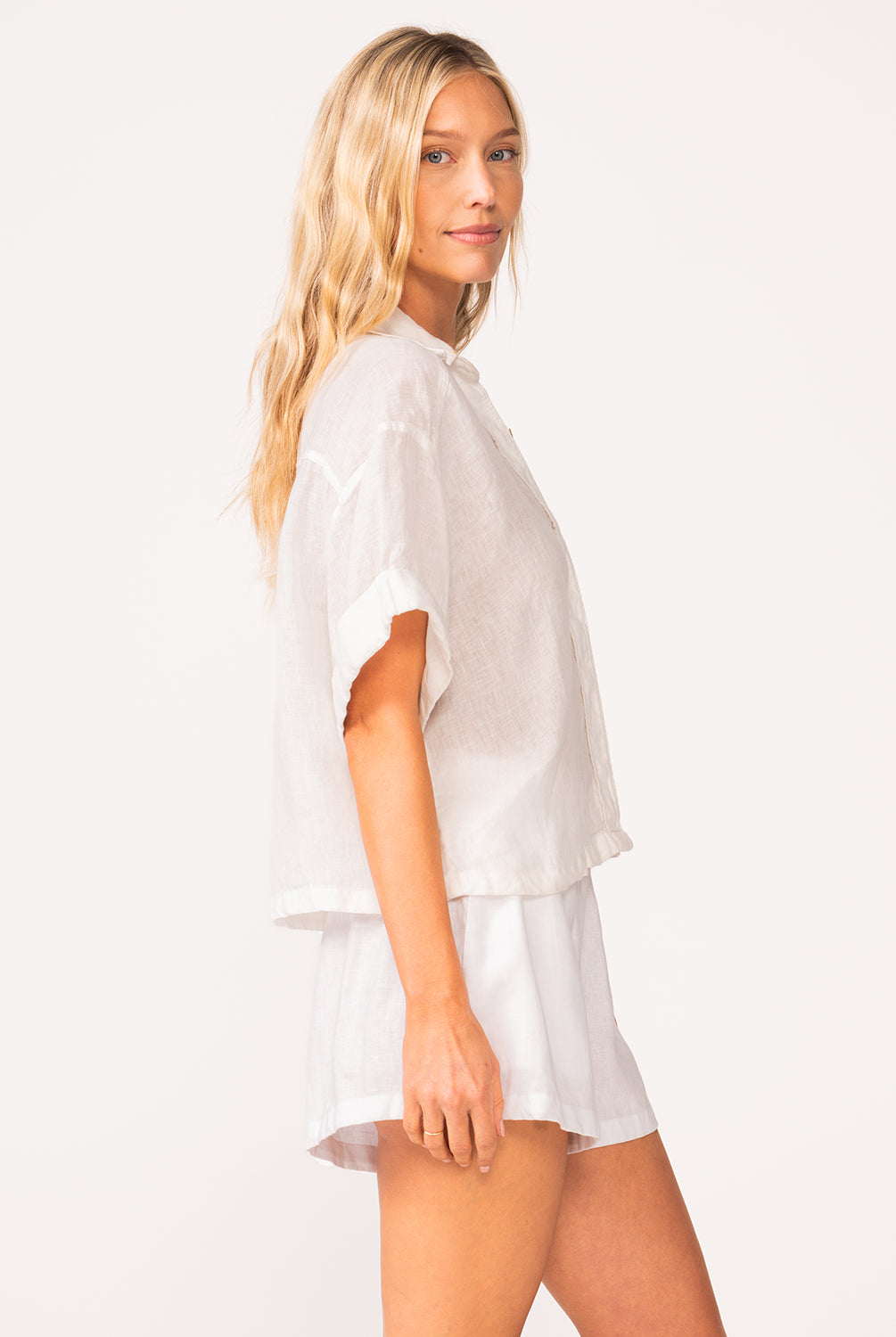A woman wearing a matching white linen shirt and short set standing against a white wall.