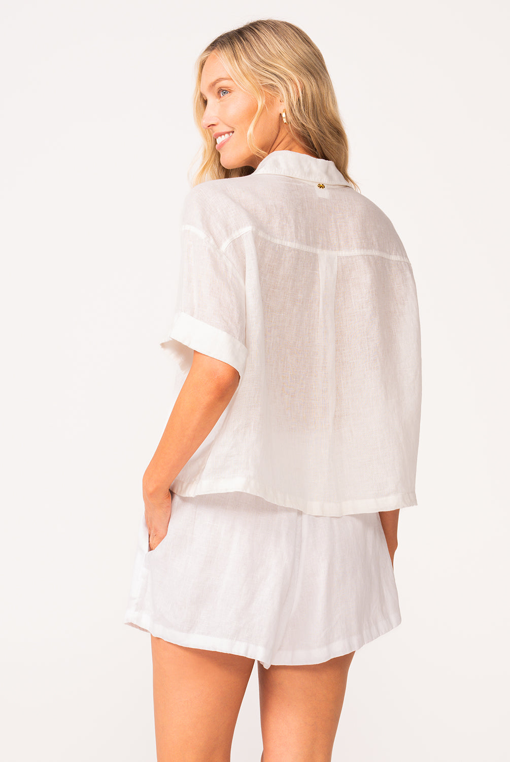 A woman wearing a matching white linen shirt and short set facing towards a white wall.