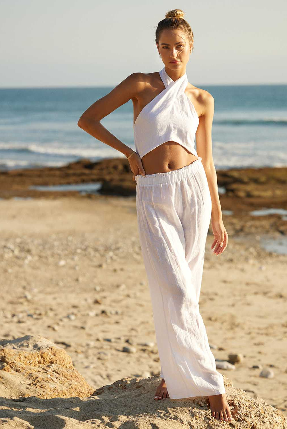 A woman wearing a white linen top and long linen pants standing on a beach near the ocean.