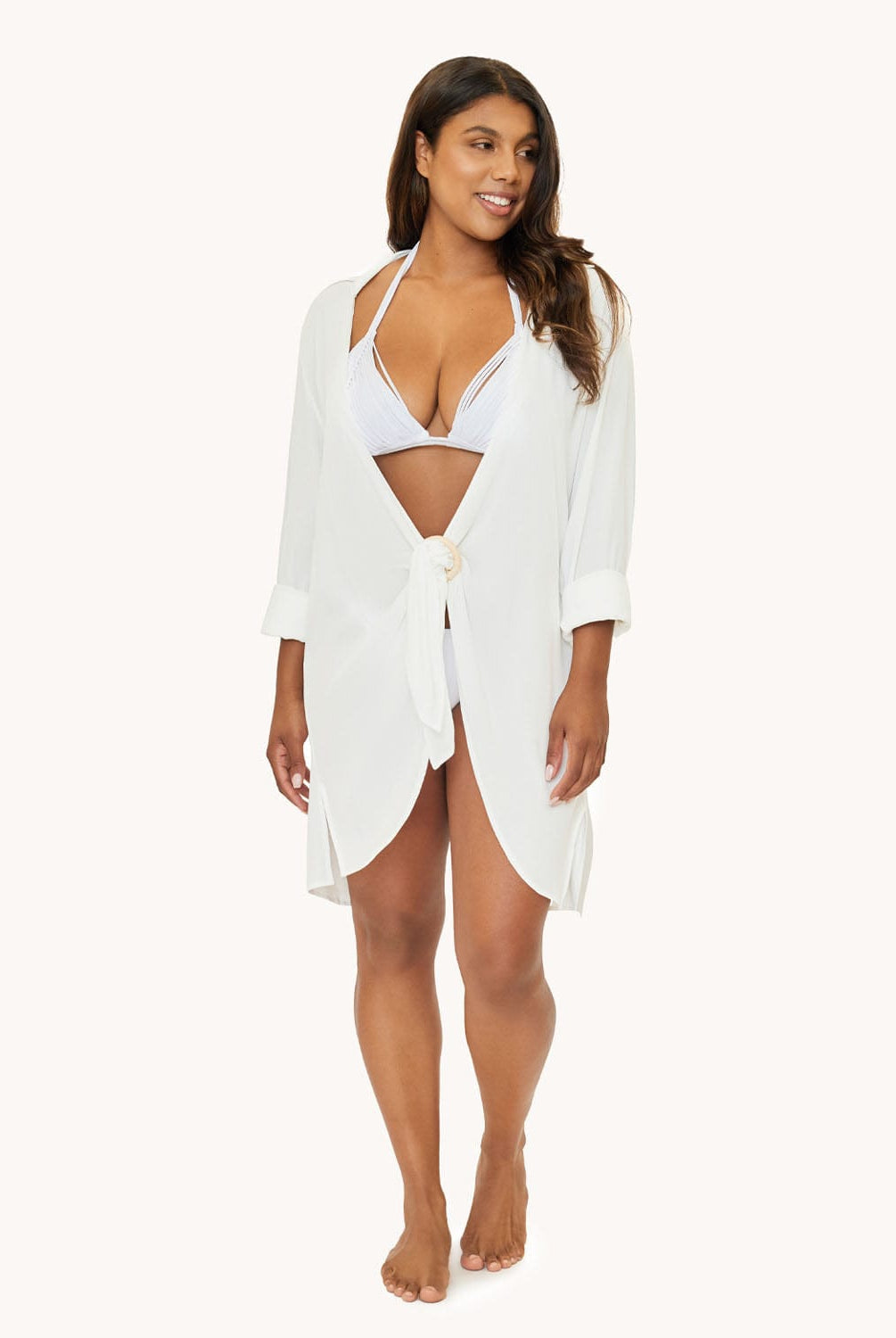 A woman wearing a white long sleeve linen cover up standing against a white wall.