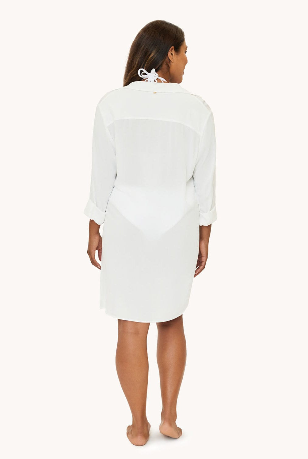 A woman wearing a white long sleeve cover up facing towards a white wall.