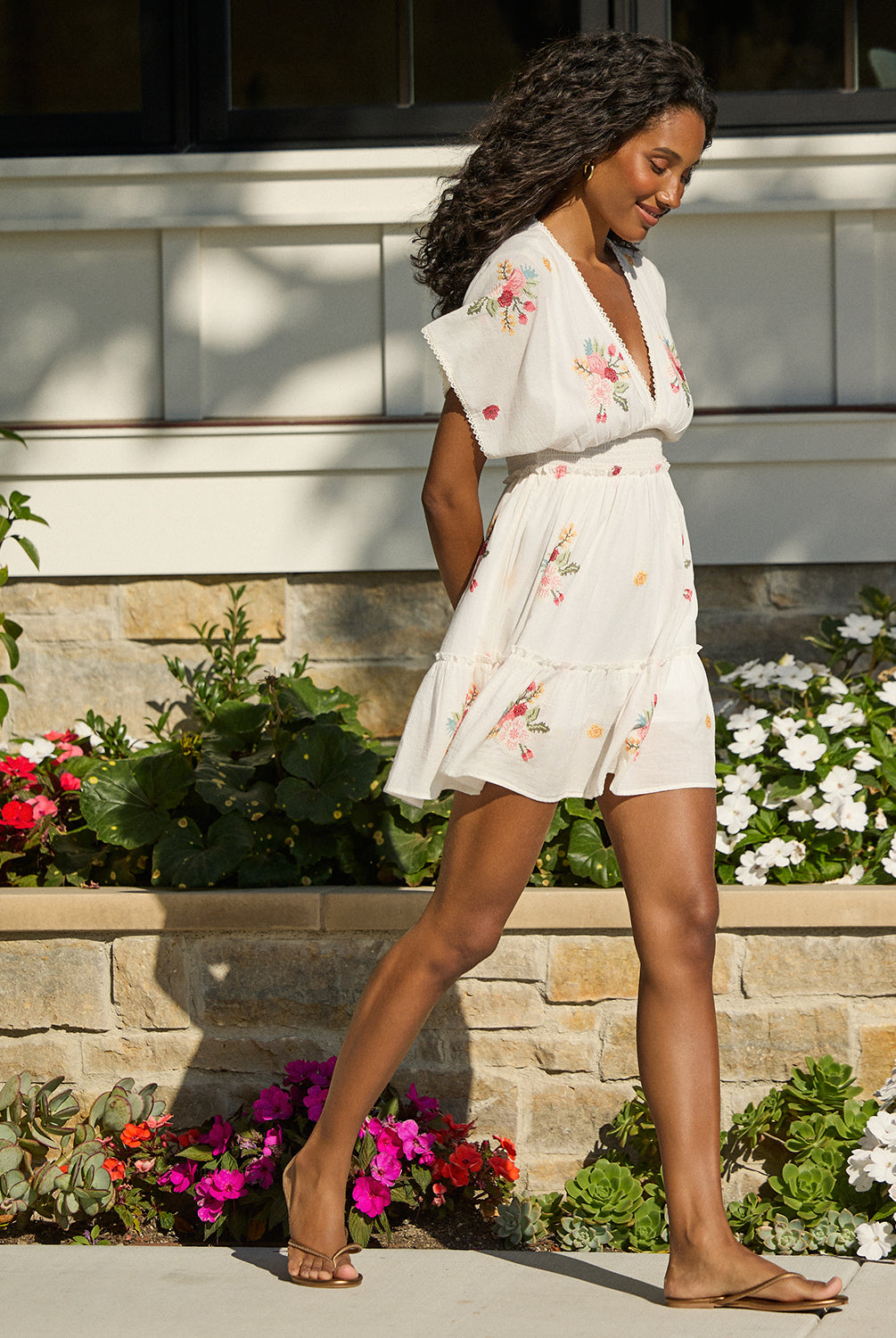 A woman wearing a short white dress with floral details walking on a sidewalk near flowers.