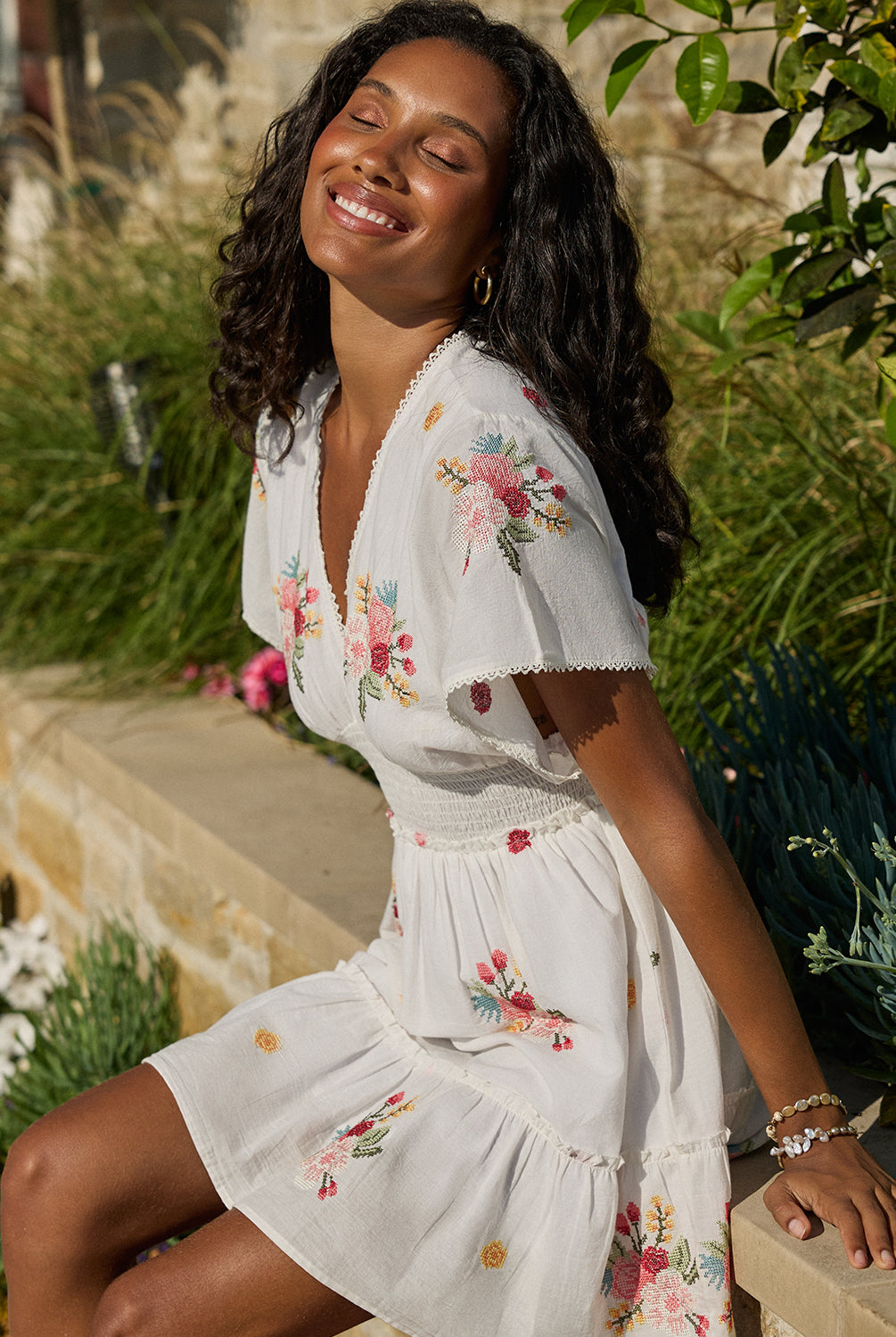 A woman wearing a short white dress with floral details sitting on a concrete ledge.
