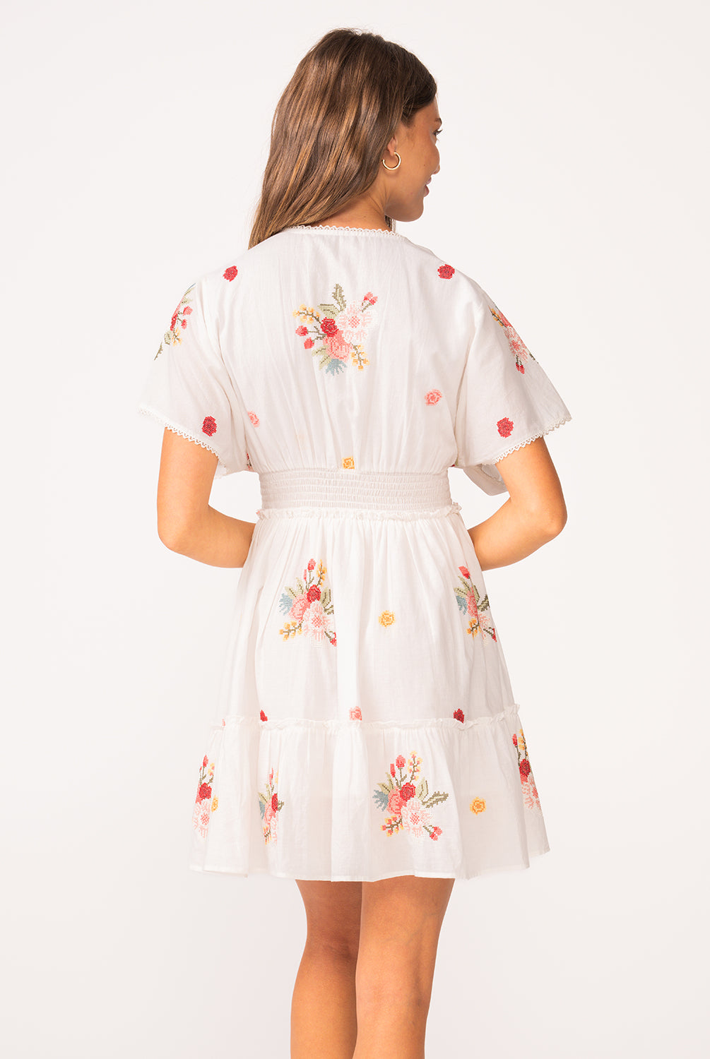A woman wearing a short sleeve dress with floral details facing towards a white wall.