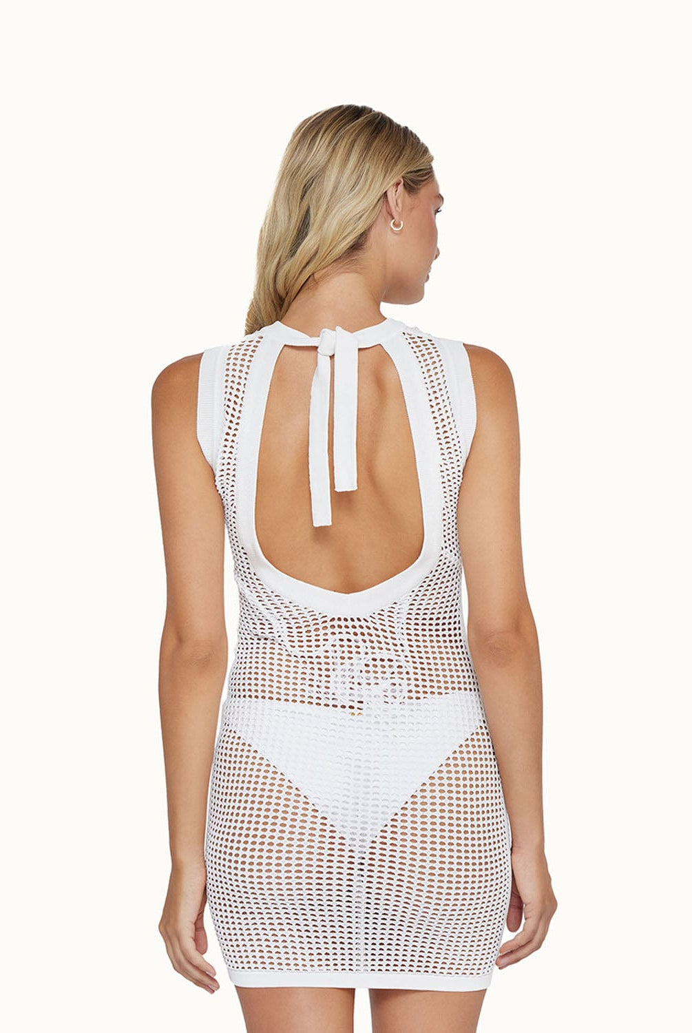 A blonde woman wearing a short white dress over a white bikini facing towards a white wall.