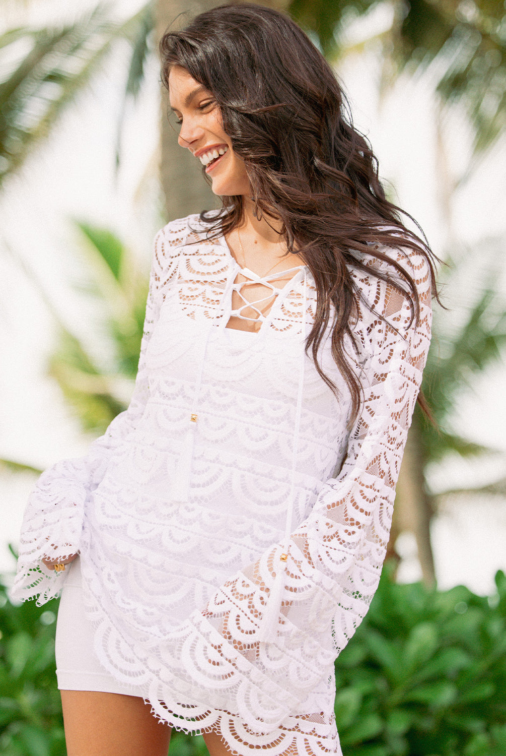 A woman wearing a white mini slip dress under a lace cover up standing anear trees outside.