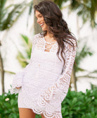 A woman wearing a white mini slip dress under a lace cover up standing anear trees outside.
