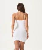 A woman wearing a white mini slip dress facing towards a white wall.