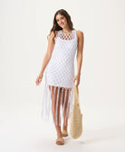 A woman wearing a white mini slip dress under a crochet cover up standing against a white wall.