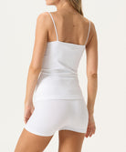 A woman wearing a white tank top and matching shorts facing towards a white wall.