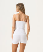 A woman wearing a white tank top and matching shorts facing towards a white wall.