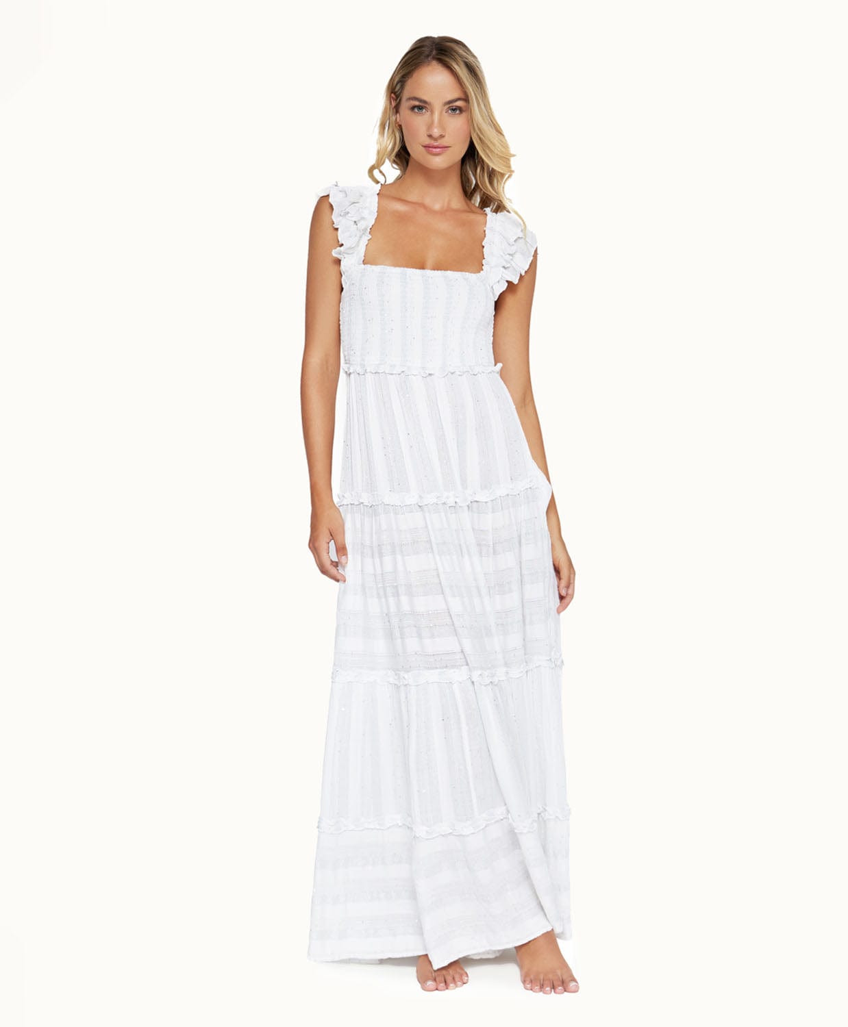 A blonde woman wearing a long white dress standing in front of a white wall.