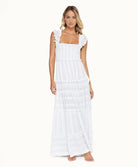 A blonde woman wearing a long white dress standing in front of a white wall.