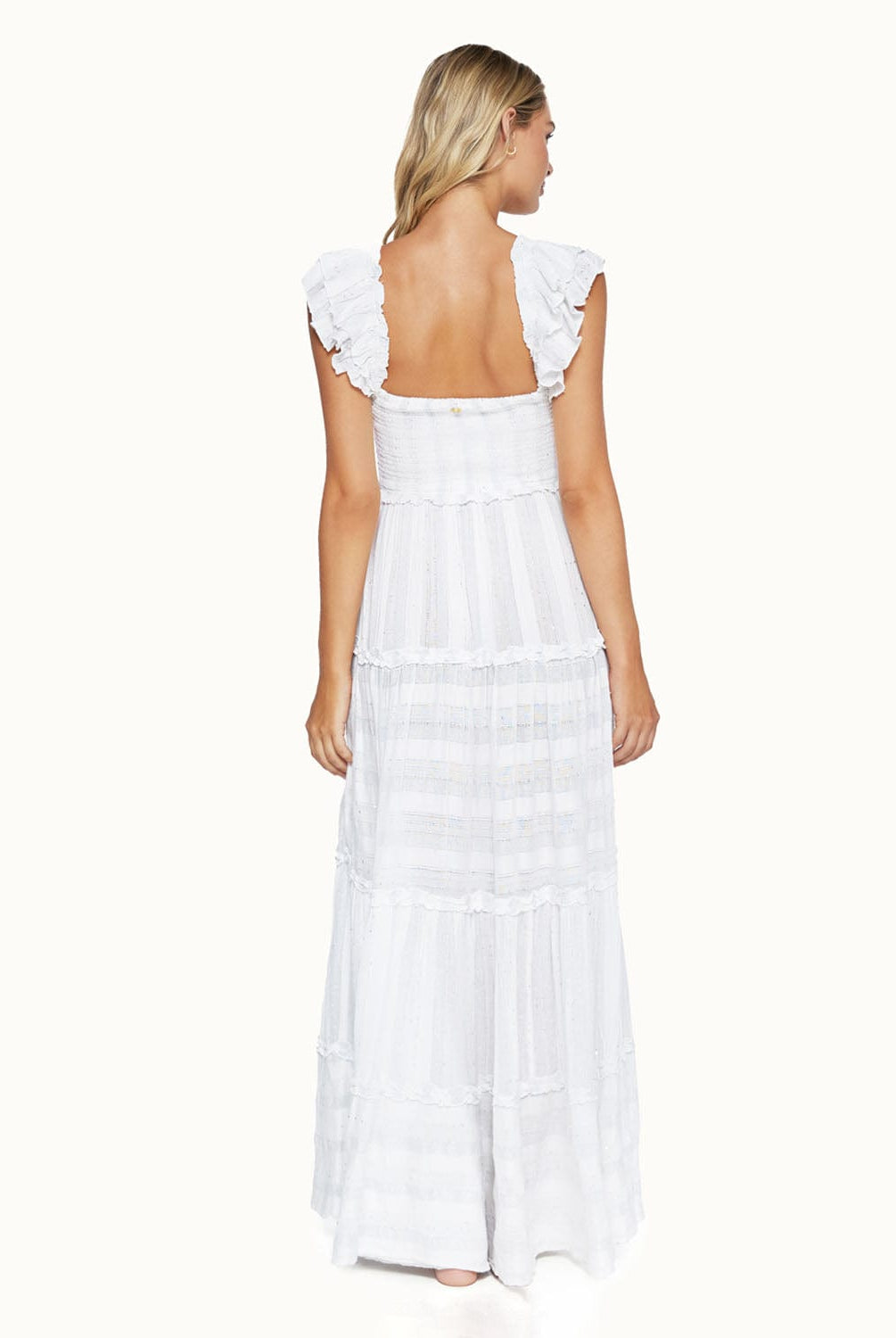 A blonde woman wearing a long white dress facing towards a white wall.