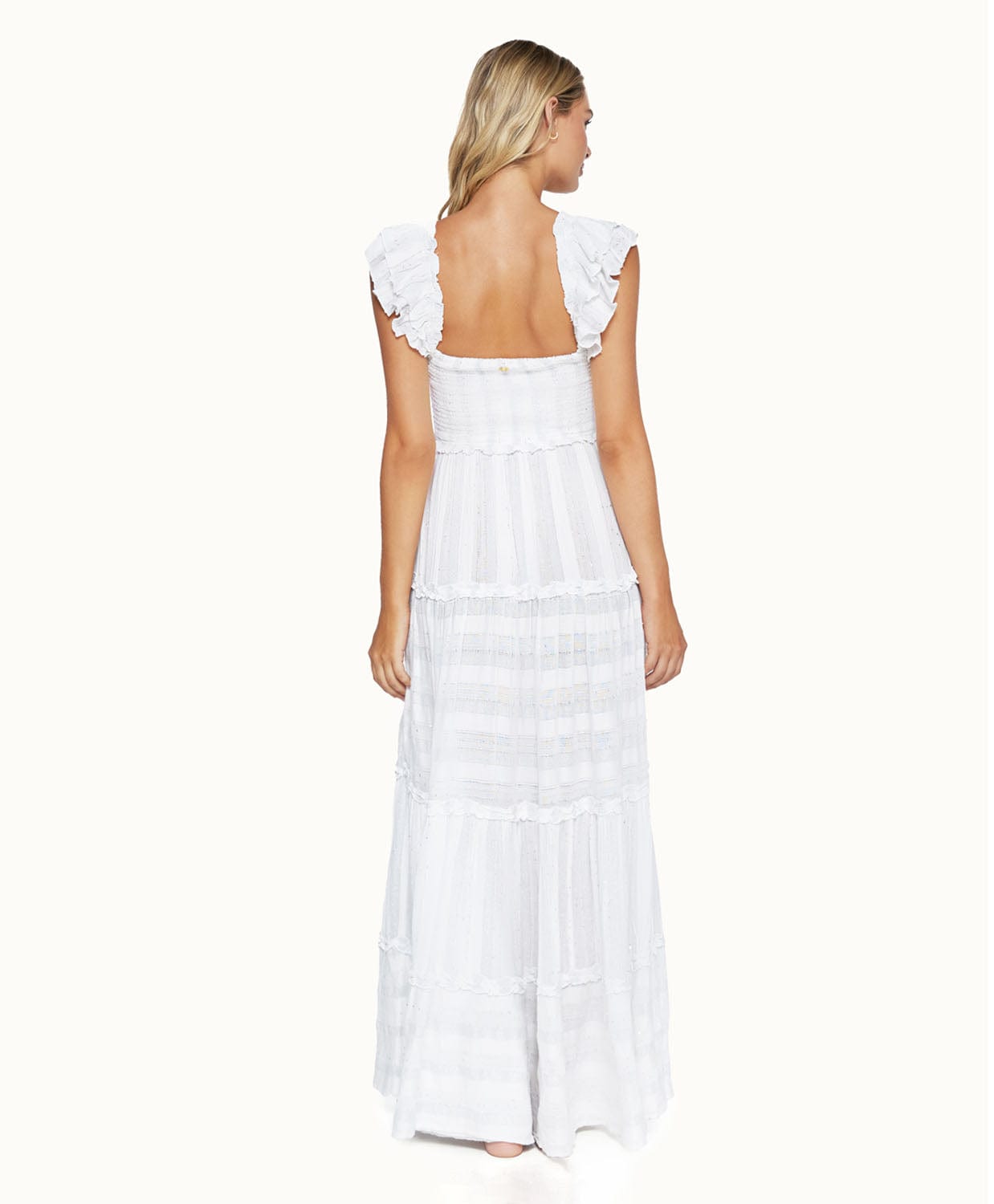 A blonde woman wearing a long white dress facing towards a white wall.