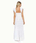 A blonde woman wearing a long white dress facing towards a white wall.