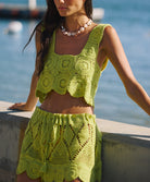 A woman wearing a matching green crochet top and skirt set leaning against a concrete wall near an ocean.
