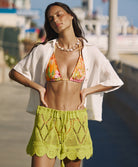 A woman wearing an orange triangle swimsuit top, a white linen shirt, and green crochet skirt. She is standing on a boardwalk near a beach.