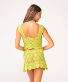 A woman wearing a matching green crochet tank and skirt set facing towards a white wall.
