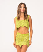 A woman wearing a matching green crochet tank and skirt set standing against a white wall.