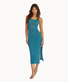 A woman wearing a teal mid-length dress standing against a white wall.