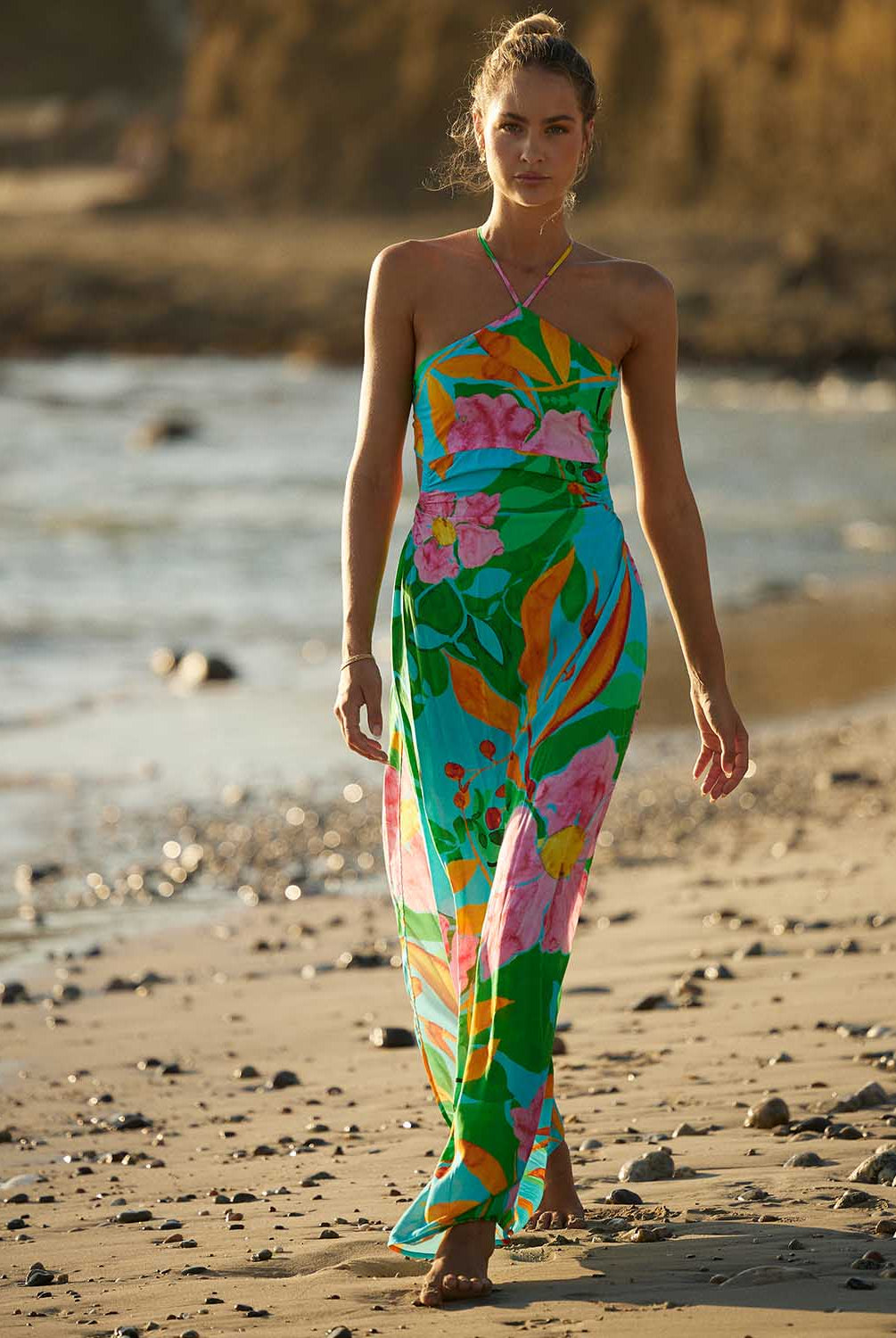 A woman wearing a long floral print dress walking on the beach near the ocean.