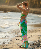 A woman wearing a long floral print dress standing on the beach near the ocean.