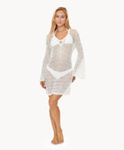 Woman wearing a white crochet bell sleeve tunic in front of a white wall.