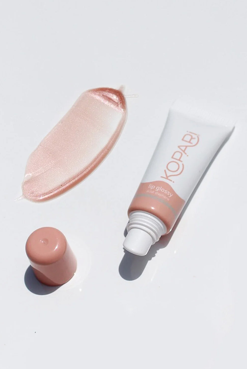 A lip gloss tube, cap, and sample against a white wall.