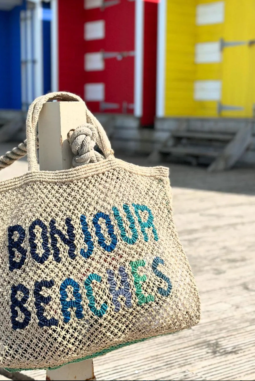 A purse that has "Bonjour Beaches" written in blue thread hung on a wooden post.
