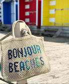 A purse that has "Bonjour Beaches" written in blue thread hung on a wooden post.