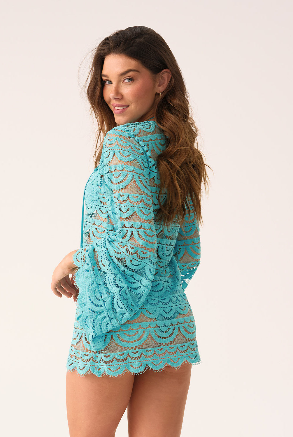 A woman in a blue lace long-sleeve cover-up smiles over her shoulder at the camera, on a white background.