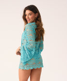 A woman in a blue lace long-sleeve cover-up smiles over her shoulder at the camera, on a white background.