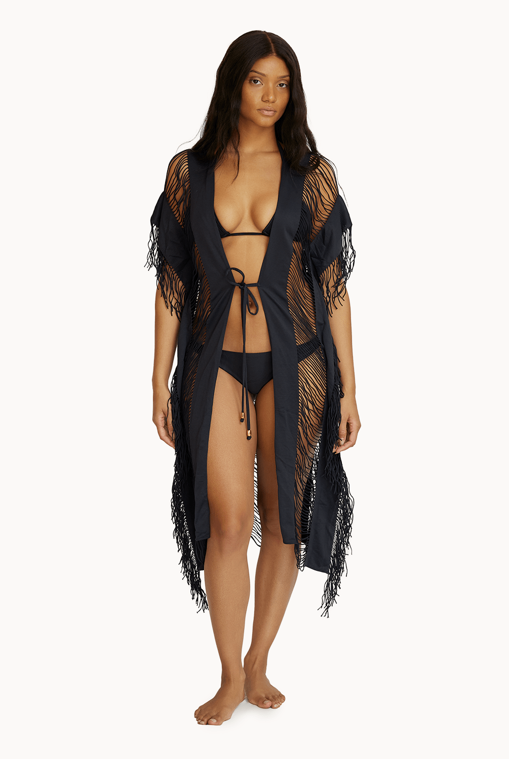 A woman wearing a black bikini and fringe cover up standing in front of a white wall.