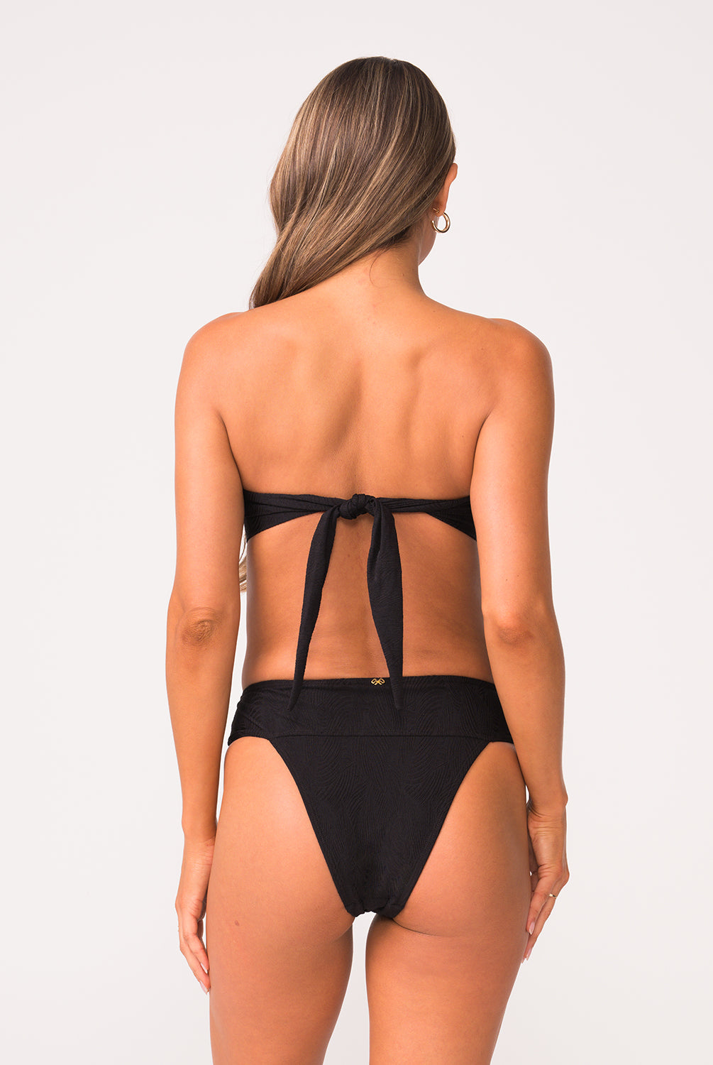 Woman wearing a black bandeau bikini top and black high waist bottom in texture fabric in front of white wall.