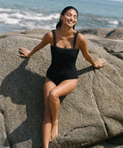 A woman sitting on a rock by the beach wearing a one-piece swimsuit with crochet trim details around the neck and shoulders.