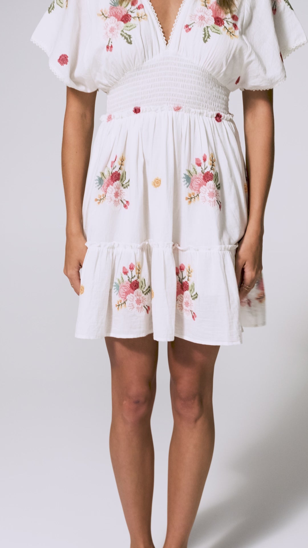 A woman wearing a short white dress with floral details spinning in front of a white wall.