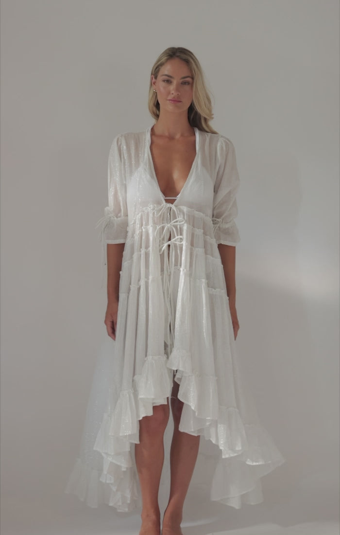 A blonde woman wearing a long white cover up dress spinning in front of a white wall. 