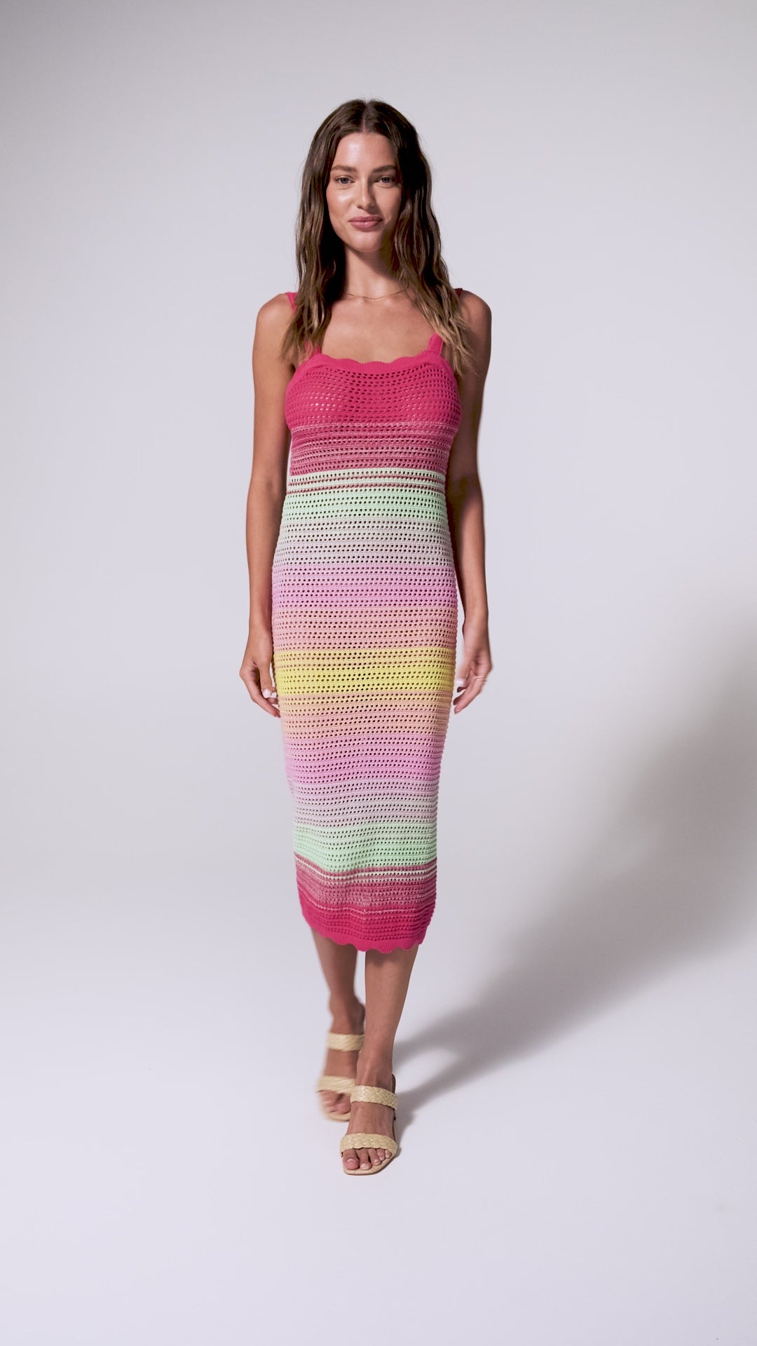 A woman wearing a pink, green, and yellow crochet knit mid length dress spinning in front of a white background.